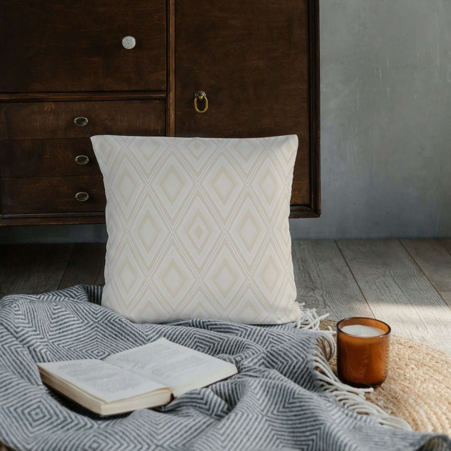 The Reid diamond LOOM Collection pillow is shown on top of a blanket along with a book and a candle.
