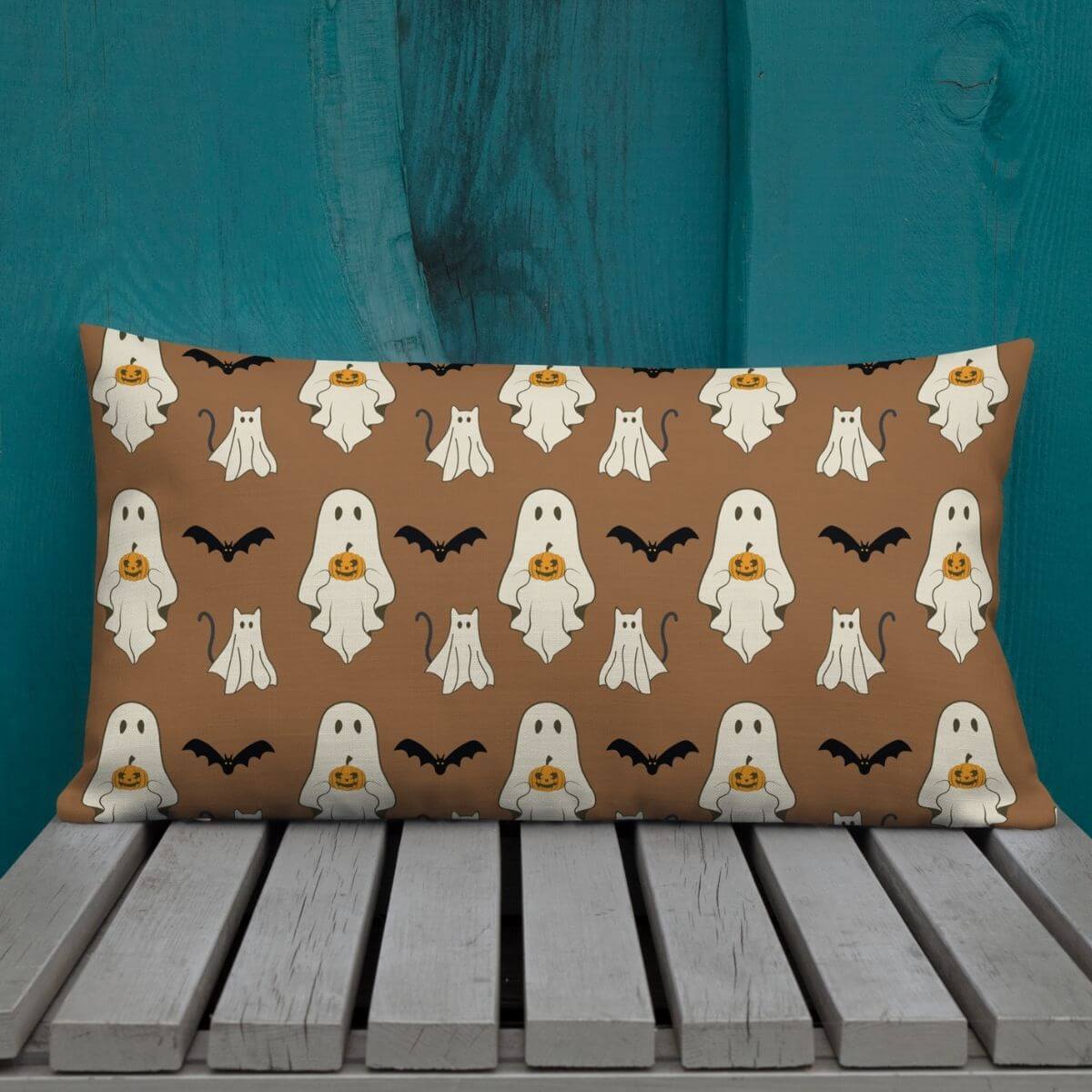 Halloween-themed pillow with ghosts, cats, and bats on a brown background, placed on a wooden surface against a teal wall.