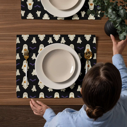 Person setting a table with Halloween-themed placemats featuring ghosts and bats.